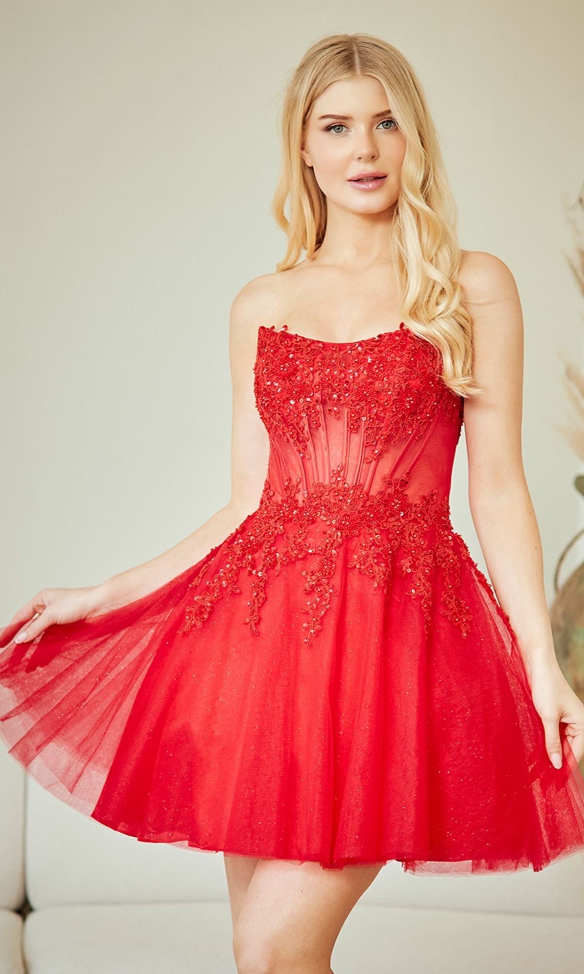 A woman wearing the Amelia Couture 9083S strapless short party dress with floral appliqué and a sheer bodice poses indoors.